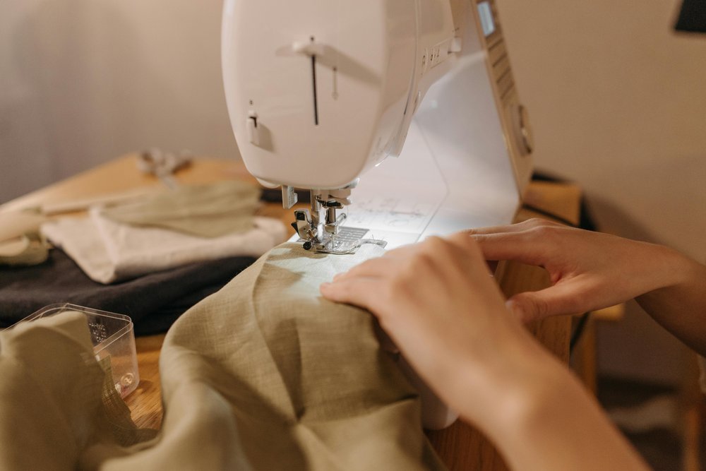 A pair of hands sewing with a white sewing machine. They are sewing a beige piece of clothing.