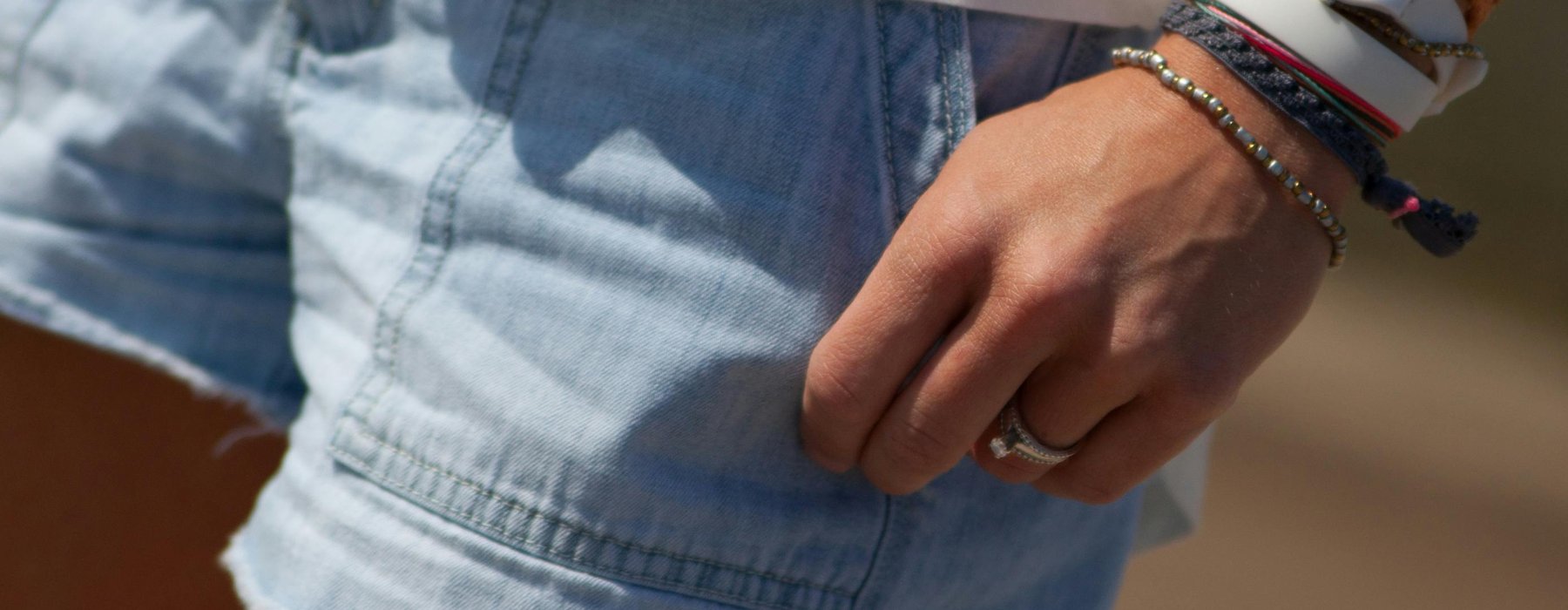 A zoomed in picture showing a pair of blue denim shorts on a person. They are also wearing a white t-shirt and on their hands you can see a watch, bands and a ring.