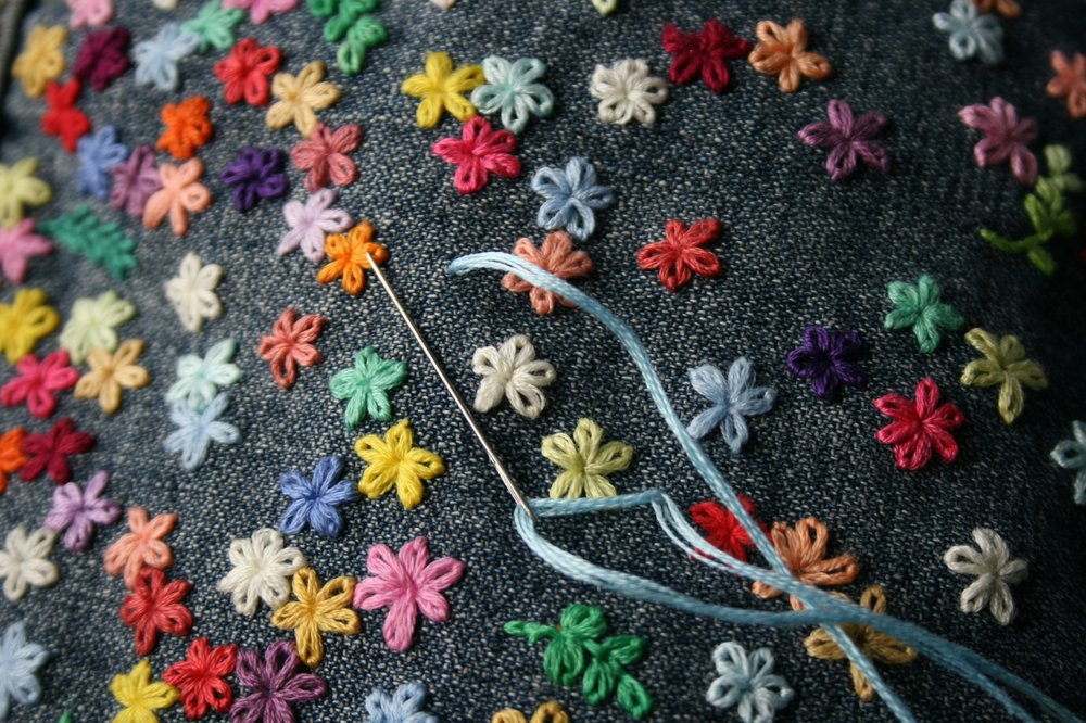 A denim piece of clothing with sewn on multi-coloured flowers with a needle and thread.