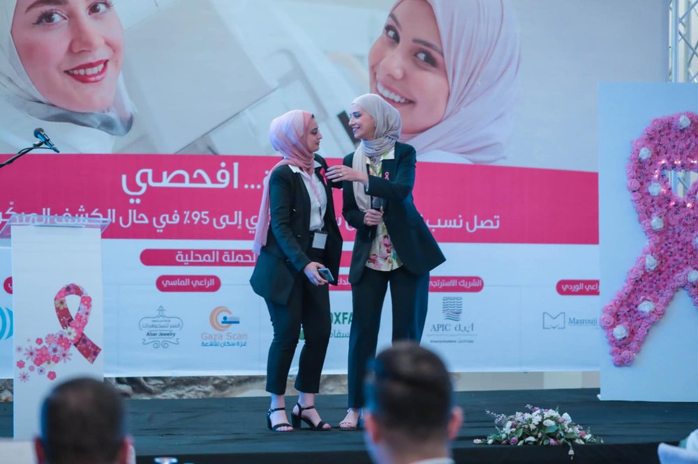Two women in head coverings stand on stage, smiling. One is holding a microphone. Behind them, a breast cancer awareness banner features a large pink ribbon.