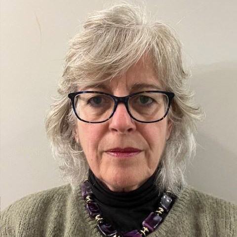 A woman wearing glasses, dressed in a green cardigan and purple statement necklace, photographed in front of a neutral background.