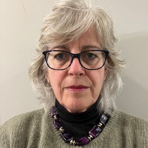 A woman wearing glasses, dressed in a green cardigan and purple statement necklace, photographed in front of a neutral background.