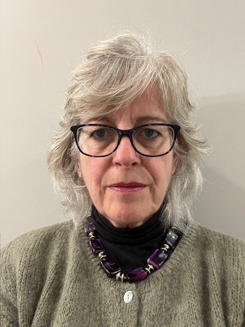 A woman wearing glasses, dressed in a green cardigan and purple statement necklace, photographed in front of a neutral background.