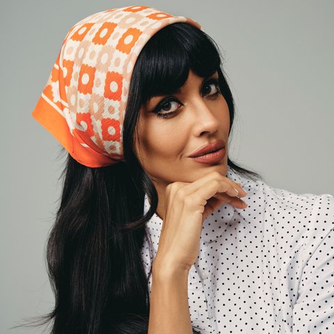 A close-up of Jameela Jamil looking into the camera. She is wearing a spotted white blouse and a floral 1960s-style bandana in her hair.
