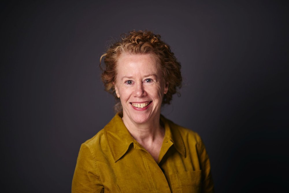 A woman with short curly red hair smiles warmly into the camera. She is wearing a mustard coloured shirt, and is posed against a plain dark grey background.
