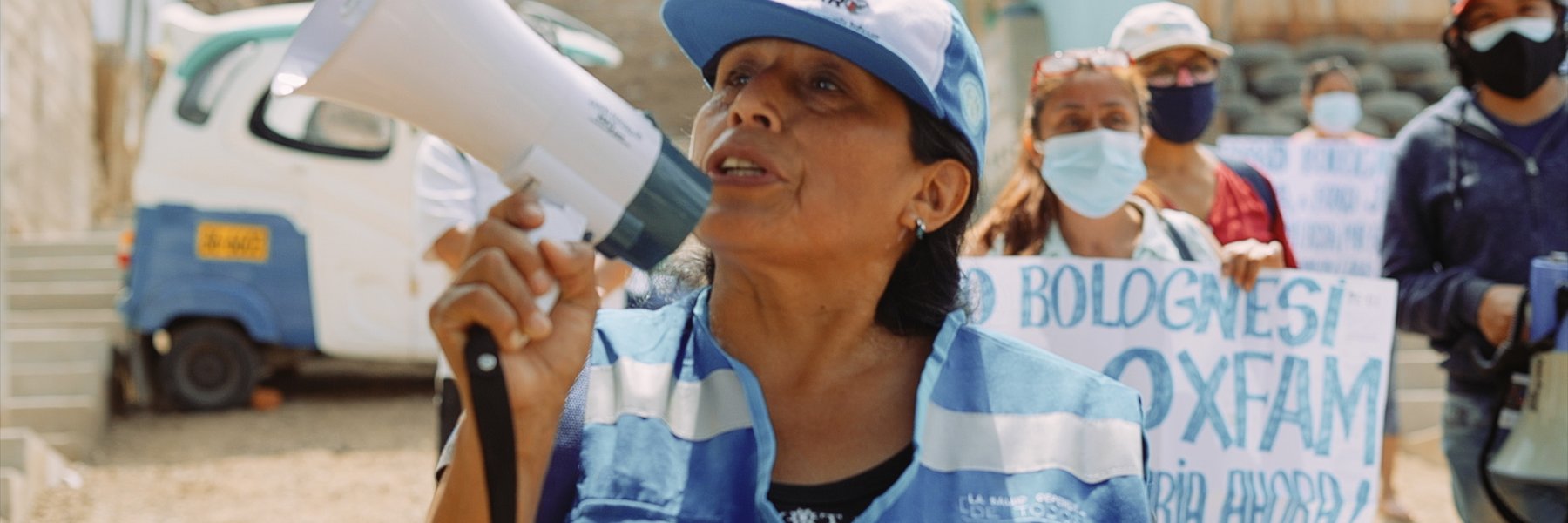 Janet wears a blue hi-vis jacket and a blue cap and carries a megaphone. Protesters stand behind her with signs.