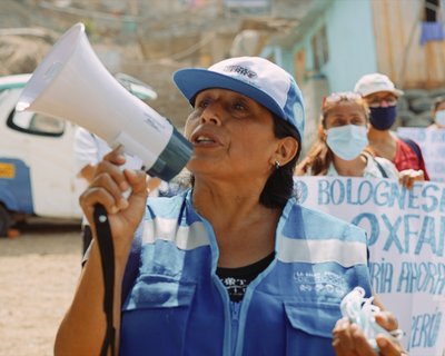 Janet wears a blue hi-vis jacket and a blue cap and carries a megaphone. Protesters stand behind her with signs.