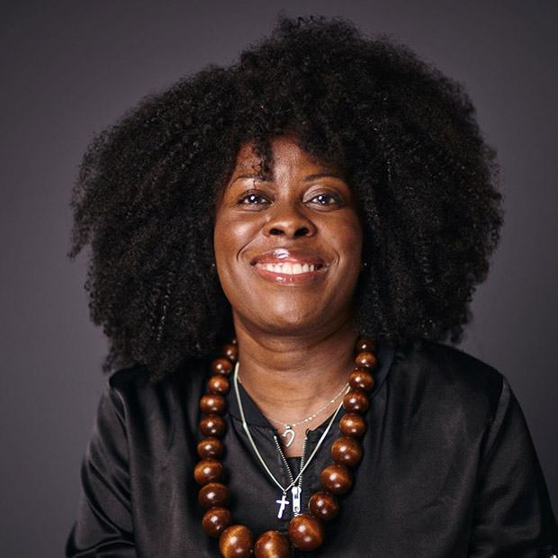 A woman with curly hair, wearing a black jacket and a large wooden bead necklace, smiles against a dark grey background.