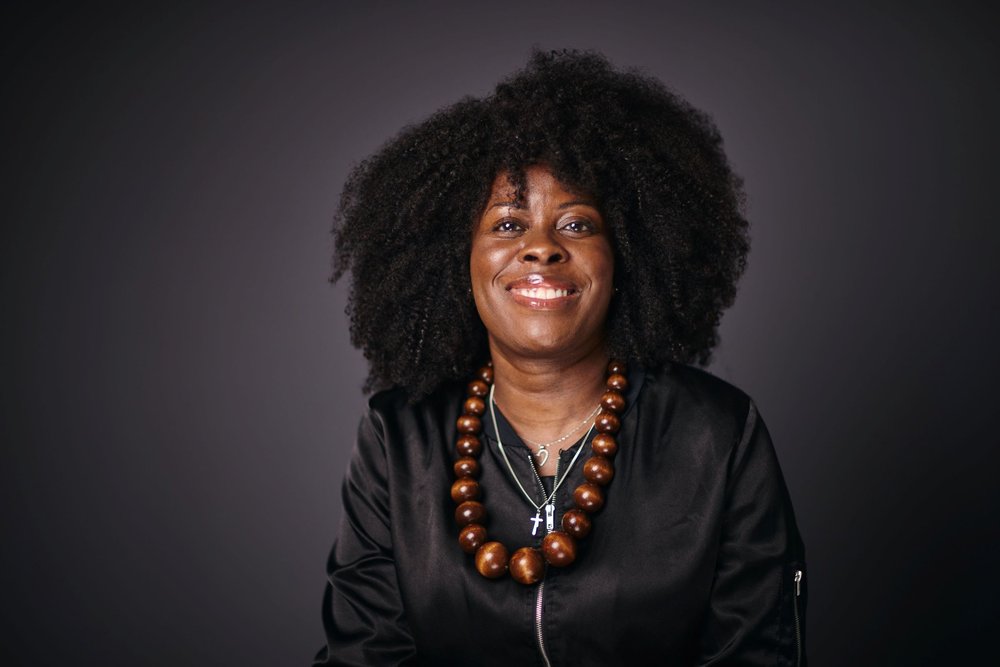 A woman with curly hair, wearing a black jacket and a large wooden bead necklace, smiles against a dark grey background.