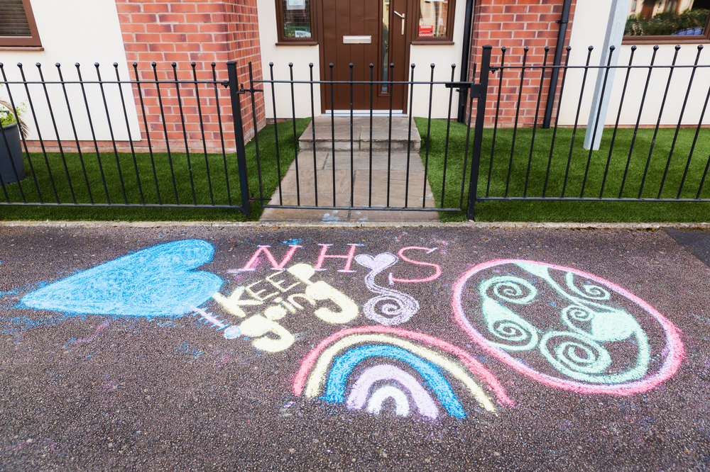 Colourful chalk on a pavement outside a 70s house with a black gate with a rainbow and the words 'keep going NHS'