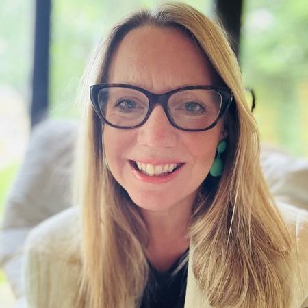 A woman with long blonde hair and black glasses smiles into the camera.