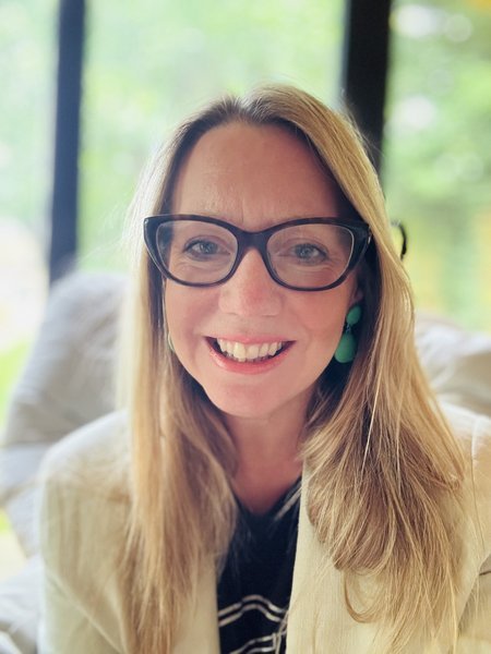 A woman with long blonde hair and black glasses smiles into the camera.