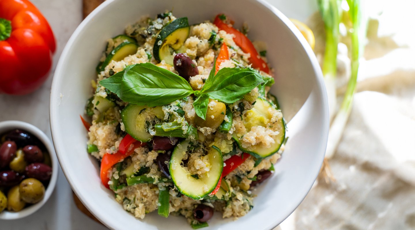 A vibrant quinoa salad featuring sliced zucchini, red bell peppers, olives, and fresh herbs is served in a white bowl, garnished with a sprig of fresh basil. A small bowl of mixed olives and a red bell pepper are visible in the background.