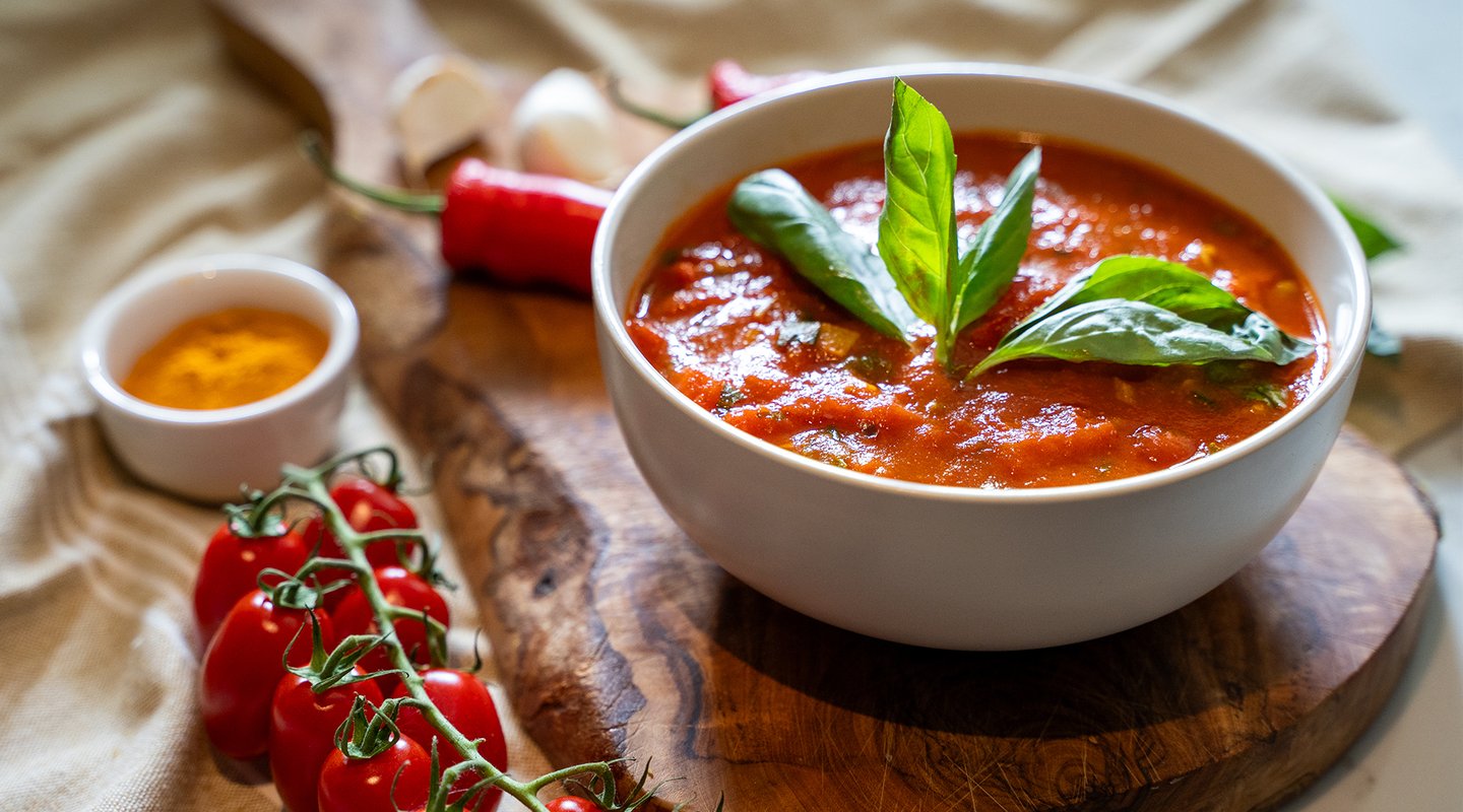 A bowl of vibrant tomato soup garnished with fresh basil leaves sits on a wooden board. Surrounding it are fresh ingredients including cherry tomatoes, red chili peppers, garlic, and a small bowl of turmeric.
