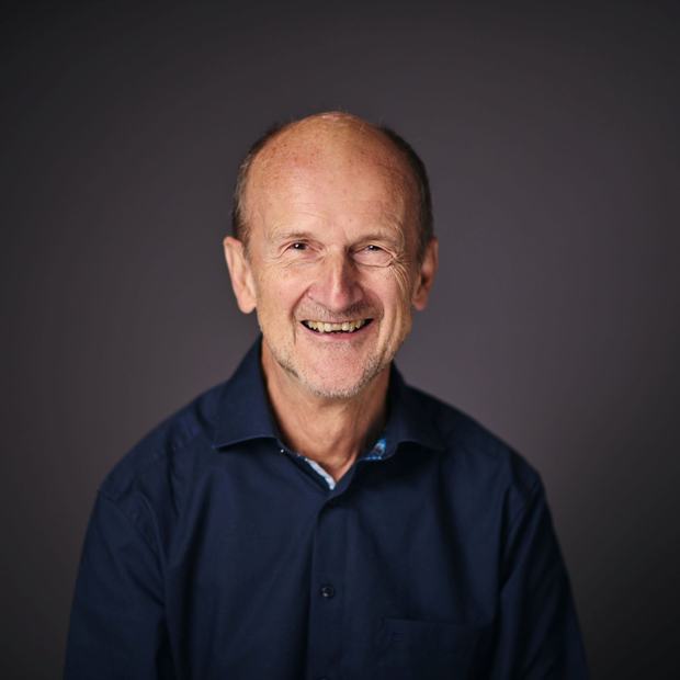 An older man wearing a dark blue dress shirt smiles into the camera, posed against a dark background.