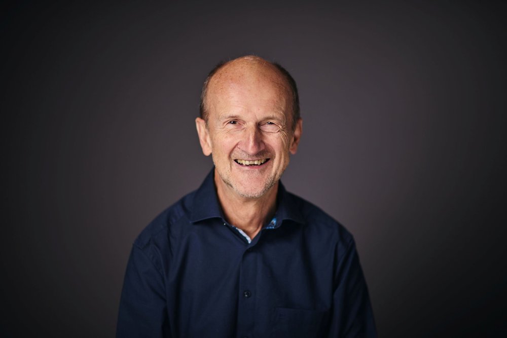 An older man wearing a dark blue dress shirt smiles into the camera, posed against a dark background.