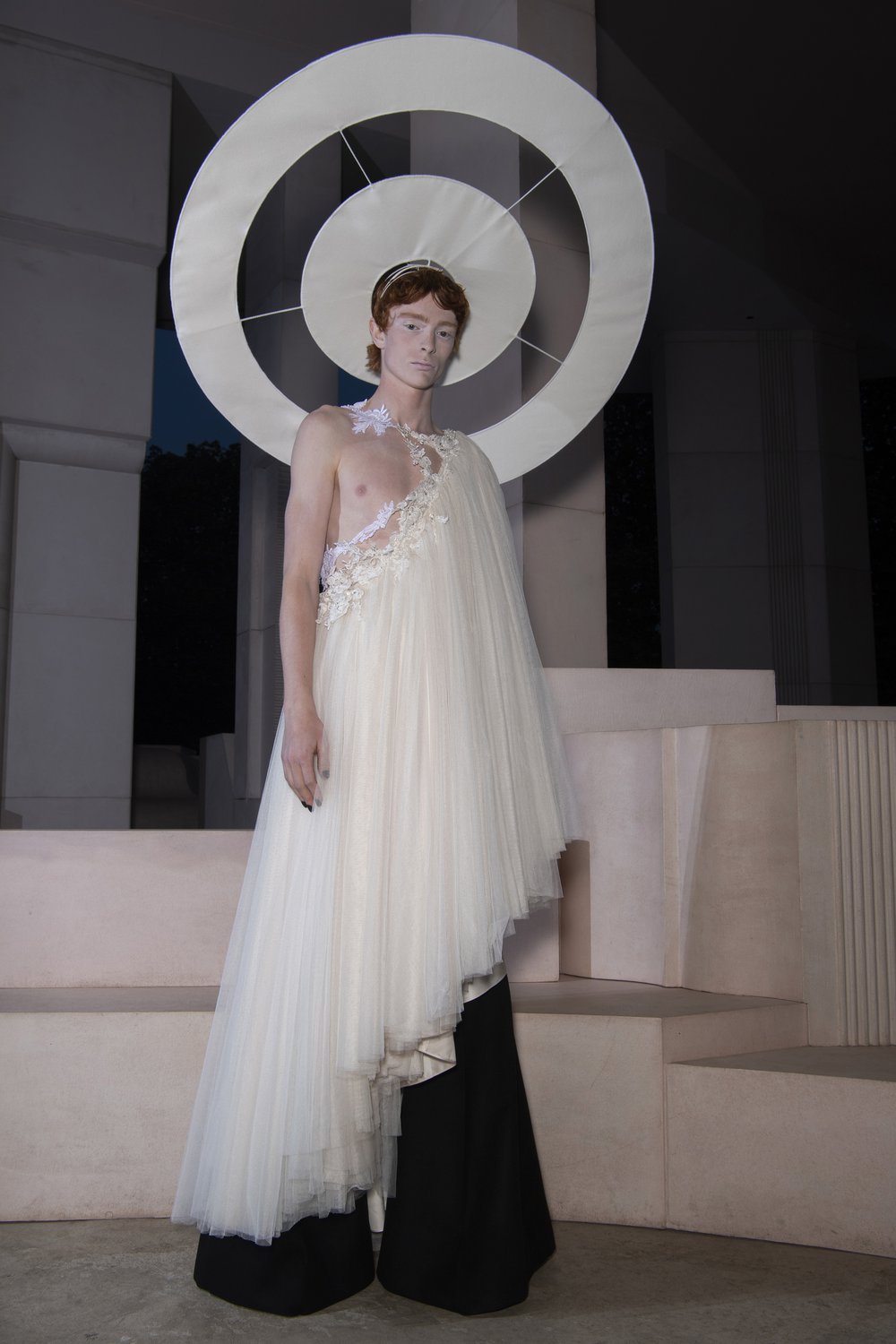 A model wears a demi-couture bridal lace toga made from Oxfam pre-loved bridal wear over black flares and a white halo.