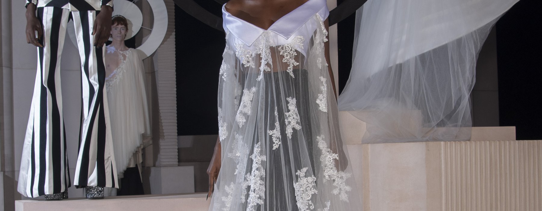 A model wears a demi-couture bridal gown made from Oxfam pre-loved bridal wear and flared suit trousers. Models stand in the background wearing other looks