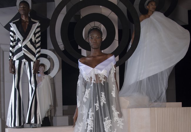 A model wears a demi-couture bridal gown made from Oxfam pre-loved bridal wear and flared suit trousers. Models stand in the background wearing other looks