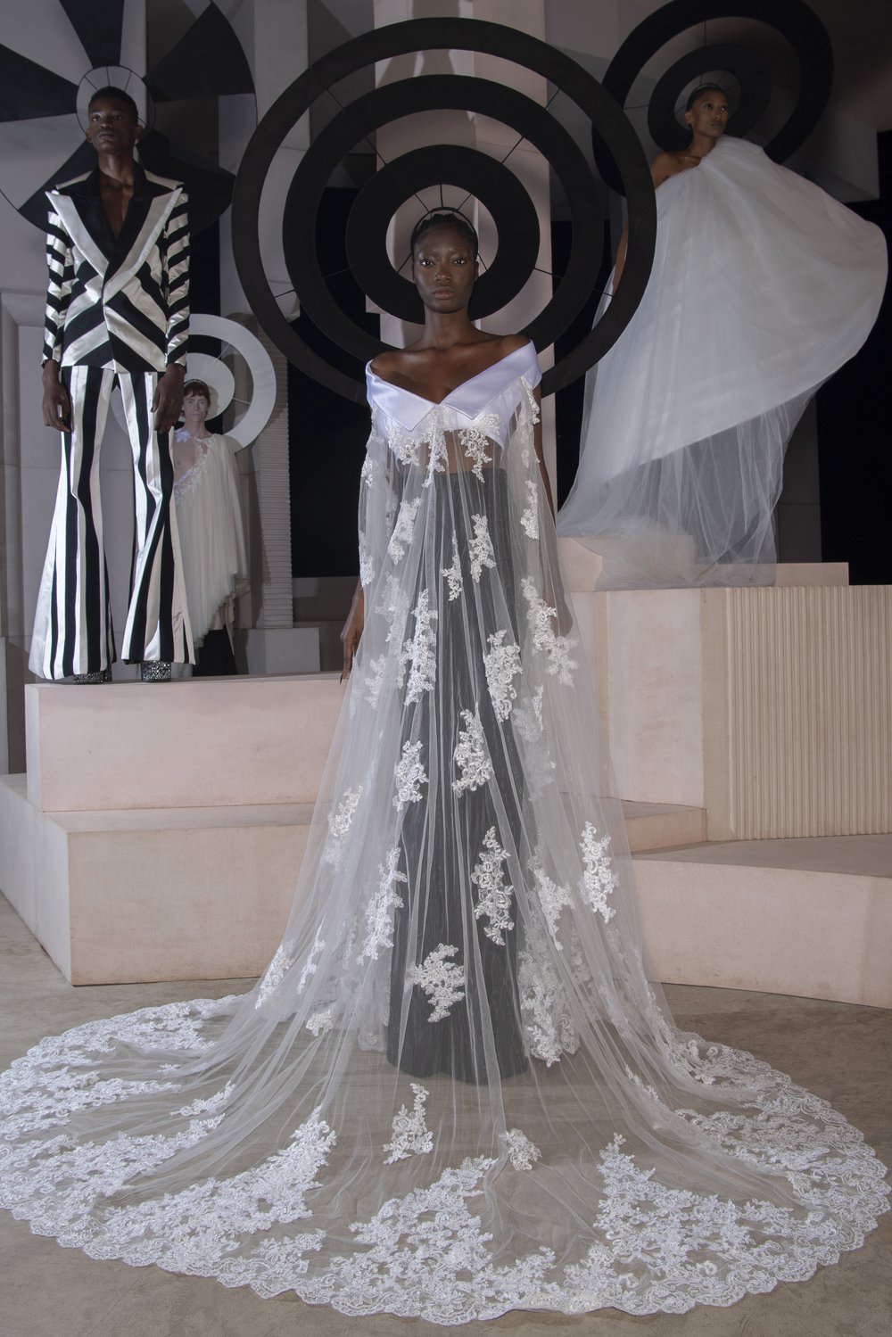 A model wears a demi-couture bridal gown made from Oxfam pre-loved bridal wear and flared suit trousers. Models stand in the background wearing other looks