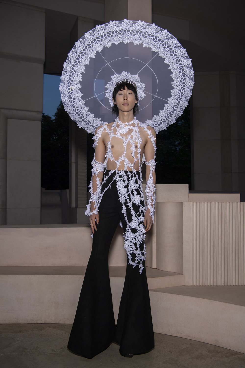 A model wears black flares and has floral detail from an Oxfam pre-loved bridal gown draped over their chest with a white halo also with the floral detail on it