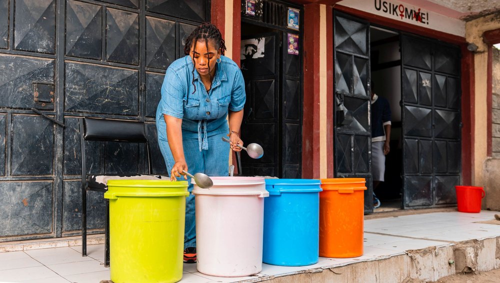 A woman in a blue denim outfit serves food from large colourful containers outside a building with a sign that reads “USIKIMYE!”.