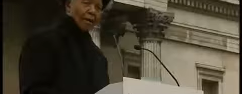 Nelson Mandela dressed in black at a podium with the logo 'Make Poverty History' giving a speech in front of a stone structure.