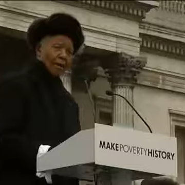 Nelson Mandela dressed in black at a podium with the logo 'Make Poverty History' giving a speech in front of a stone structure.
