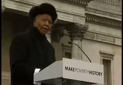 Nelson Mandela dressed in black at a podium with the logo 'Make Poverty History' giving a speech in front of a stone structure.