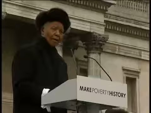Nelson Mandela dressed in black at a podium with the logo 'Make Poverty History' giving a speech in front of a stone structure.