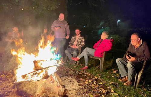 People sitting around a large outdoor bonfire at night, with chairs on grass and fallen leaves scattered on the ground.