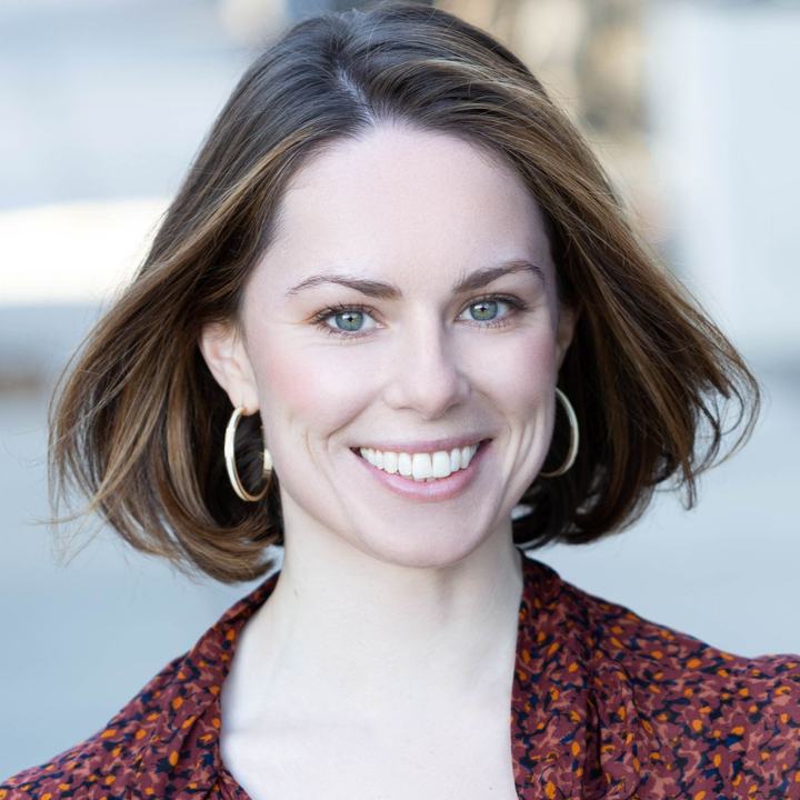 A woman with bobbed brown hair smiles into the camera. She is wearing a printed top and gold hoop earrings.