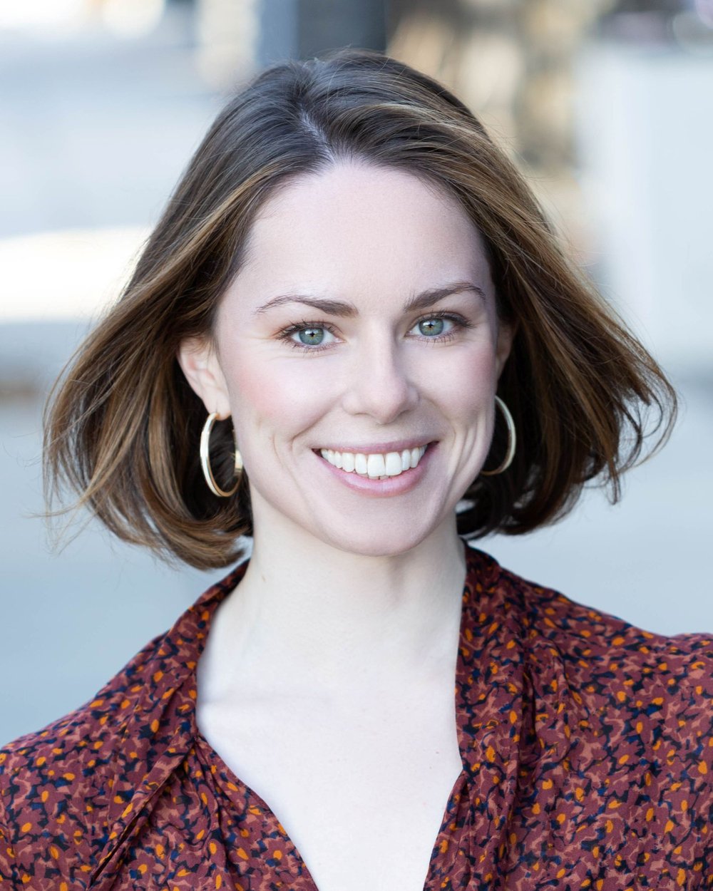 A woman with bobbed brown hair smiles into the camera. She is wearing a printed top and gold hoop earrings.