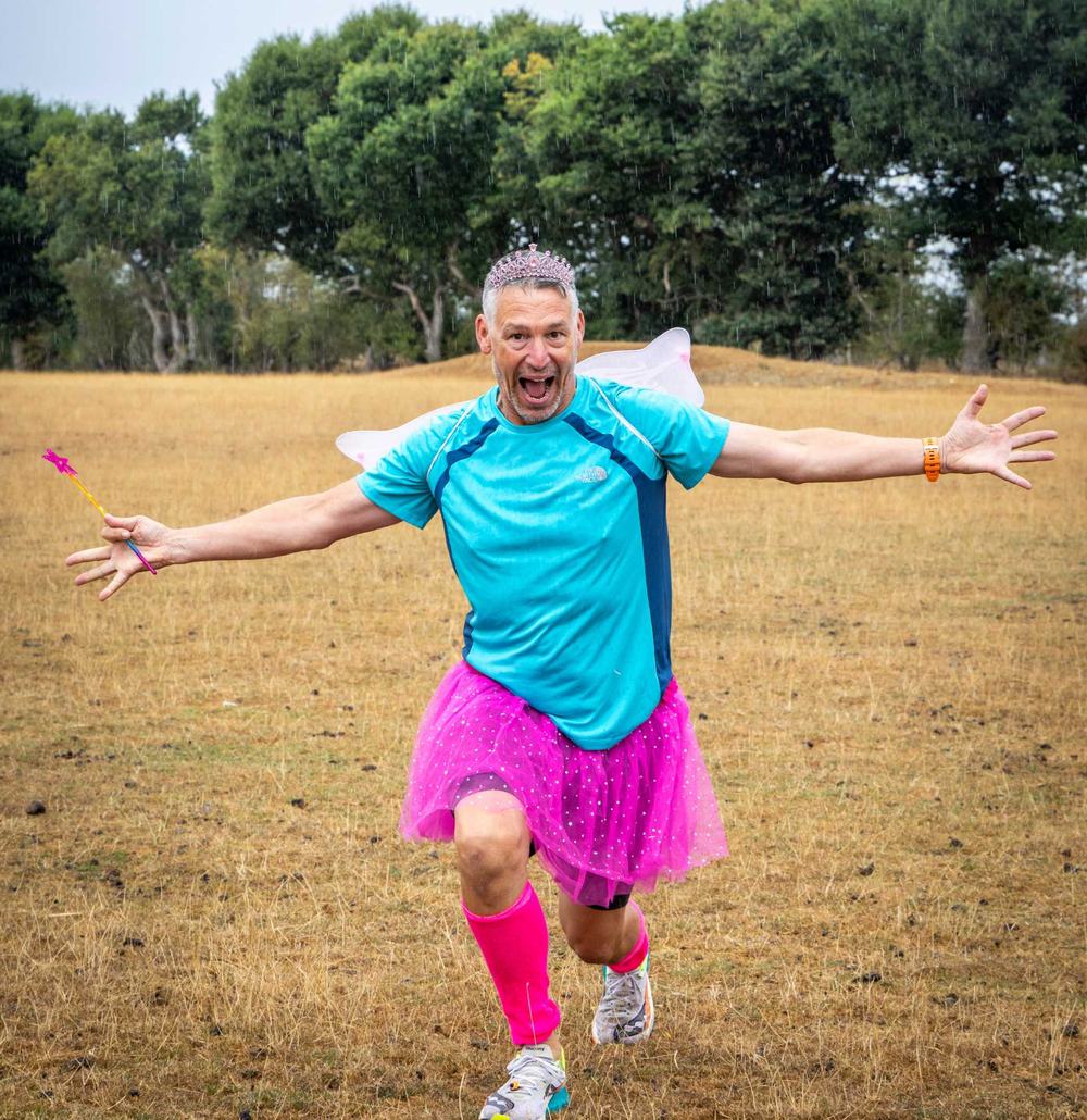 Matt is running towards the camera in a field with his arms outstretched. He is wearing a pink tutu and socks with a blue tshirt and fairy wings.