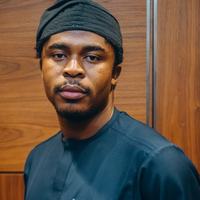 Maxwell Osasere Osarenkhoe from Oxfam in Nigeria posing for a headshot, stood in front of a wood-panelled wall, wearing a dark navy blue tunic and a black hat.