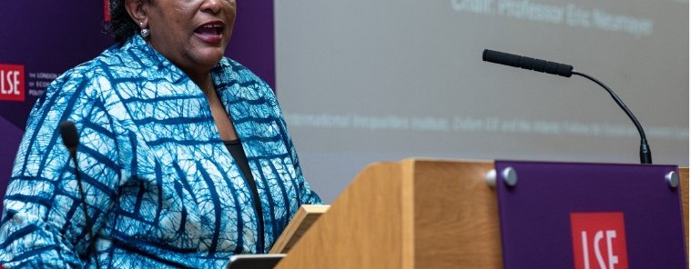 Mia Mottley standing at a wooden podium with a large screen behind her giving a speech to the London School of Economics.