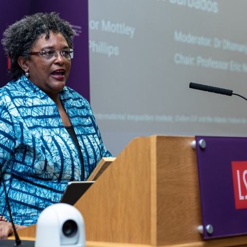 Mia Mottley standing at a wooden podium with a large screen behind her giving a speech to the London School of Economics.