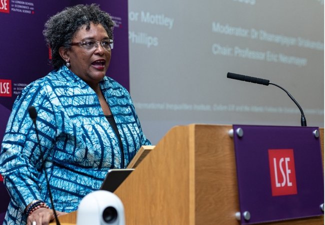 Mia Mottley standing at a wooden podium with a large screen behind her giving a speech to the London School of Economics.