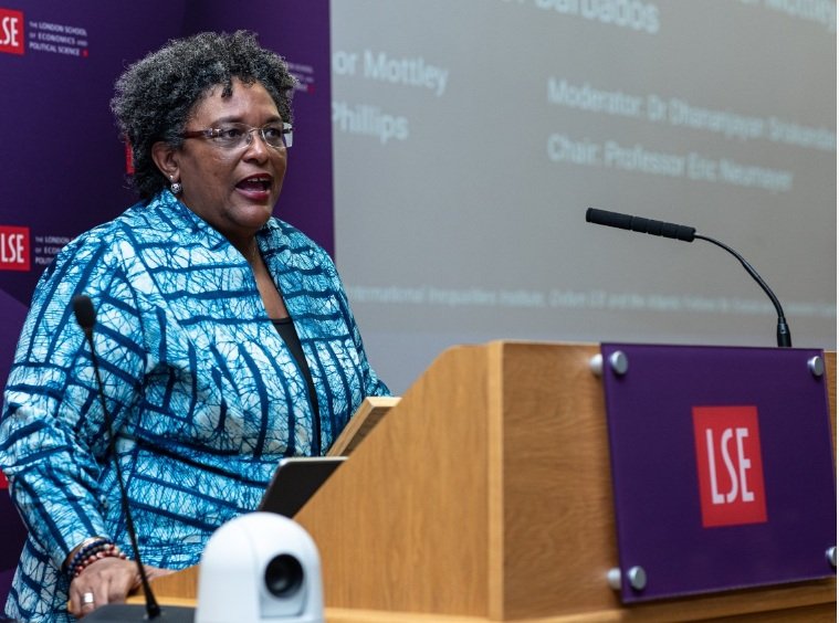 Mia Mottley standing at a wooden podium with a large screen behind her giving a speech to the London School of Economics.