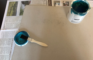 A thin chipboard or cardboard rectangle qith an open pot of blue paint and a paintbrush