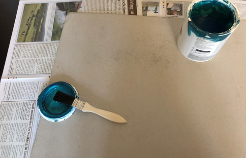 A thin chipboard or cardboard rectangle qith an open pot of blue paint and a paintbrush