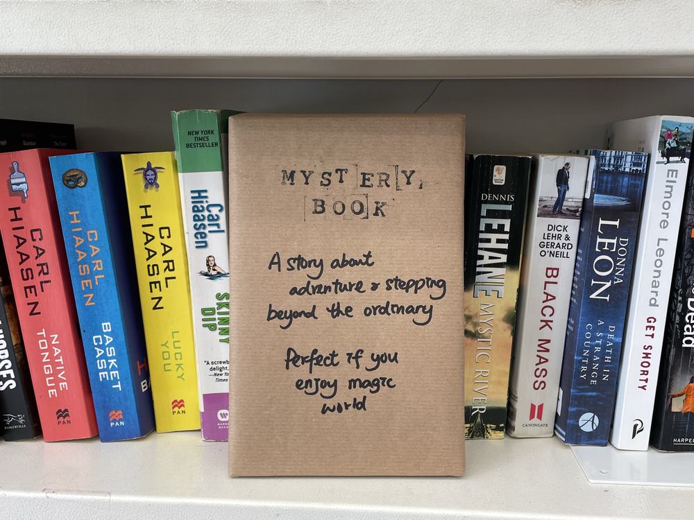 A wrapped "mystery book" displayed on a bookshelf among novels, with a handwritten note describing it as an adventure story that steps beyond the ordinary and is perfect for readers who enjoy magic.