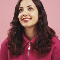 A woman with long dark wavy hair smiles up and away from the camera. She wears a bright pink jumper and red lipstick, and is photographed against a pink background.