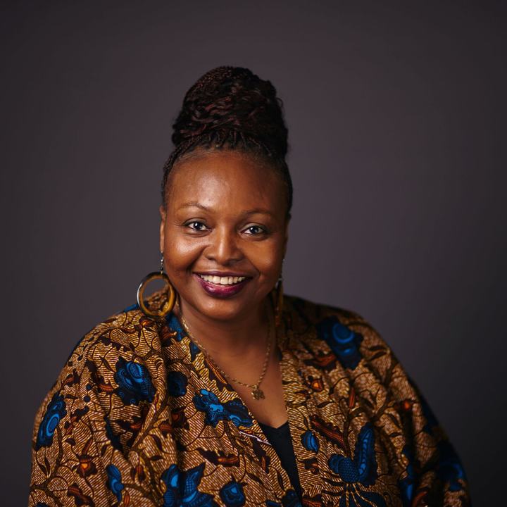 A woman wearing a blue and brown printed top and large gold earrings smiles into the camera, posed against a dark background.