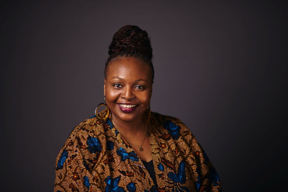 A woman wearing a blue and brown printed top and large gold earrings smiles into the camera, posed against a dark background.
