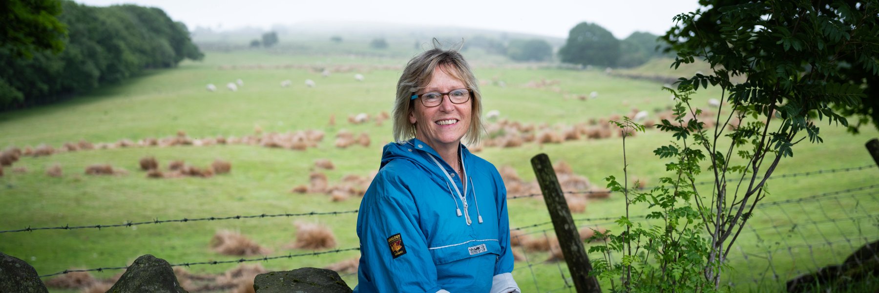 Nicky, Legacy pledger and Oxfam supporter, sits smiling on a dry stone wall.