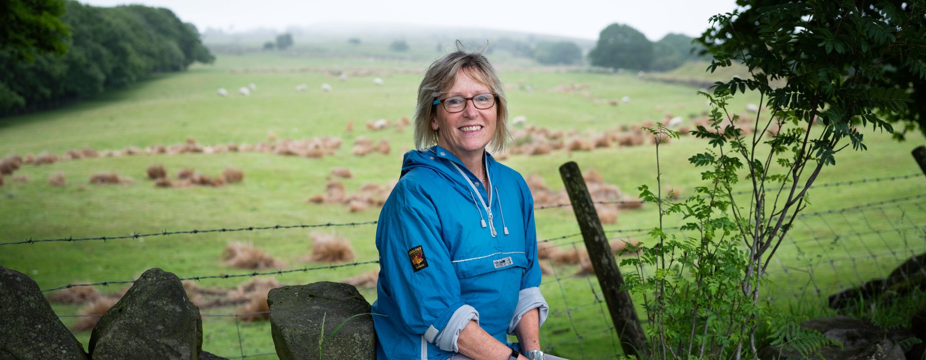 Nicky, Legacy pledger and Oxfam supporter, sits smiling on a dry stone wall.