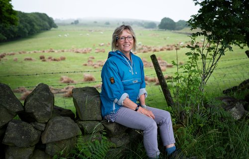 Nicky, Legacy pledger and Oxfam supporter, sits smiling on a dry stone wall.