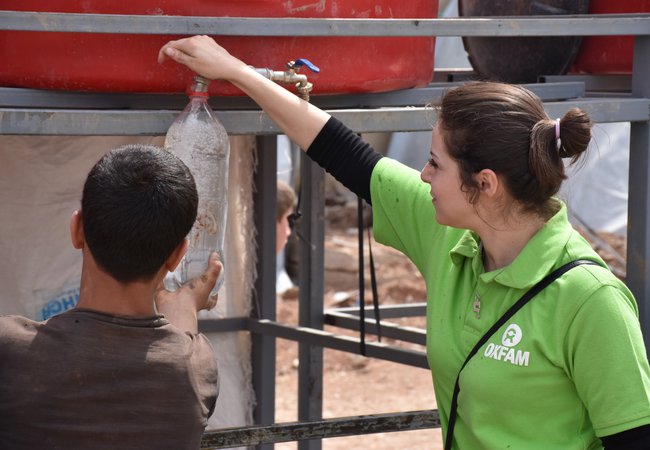 An Oxfam staff member helps a child fill a water bottle from a tank in Herjalleh collective shelter.