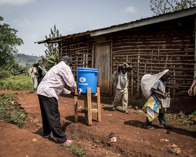 A man washes his hands under a bucket with a tap outside as people walk past.